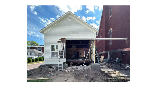 White house under construction with removed facade, ladders, and exposed framework. Brick building adjacent.