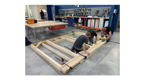 People assembling a wooden frame in a workshop with tools on the walls.