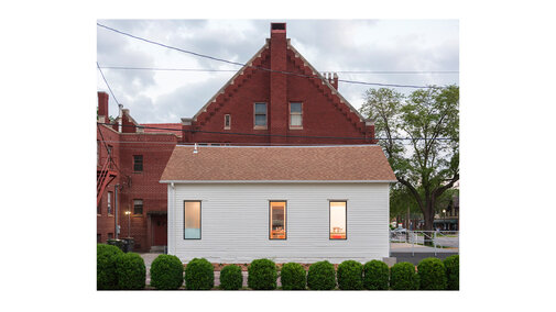 White house with glowing windows, red brick building in background, greenery in the foreground.