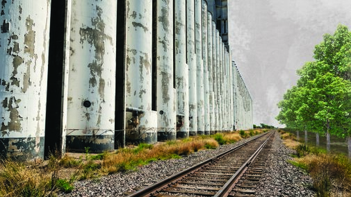 Weathered silos beside a railway track, surrounded by greenery under a cloudy sky.