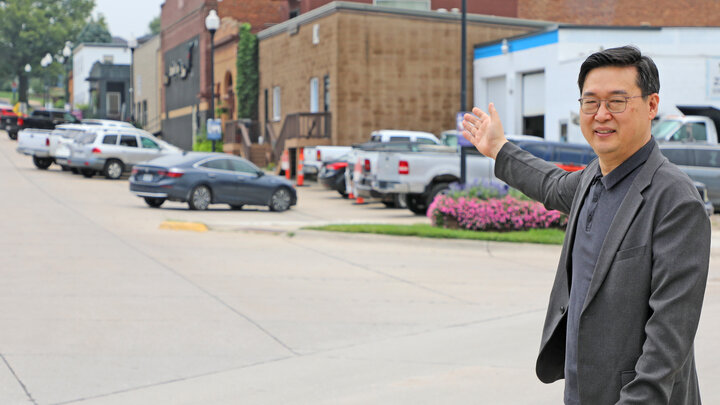 Man in a blazer gestures towards a street lined with parked cars and brick buildings.