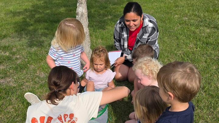 Adults and young children sitting on grass outside, engaged together in a group activity.