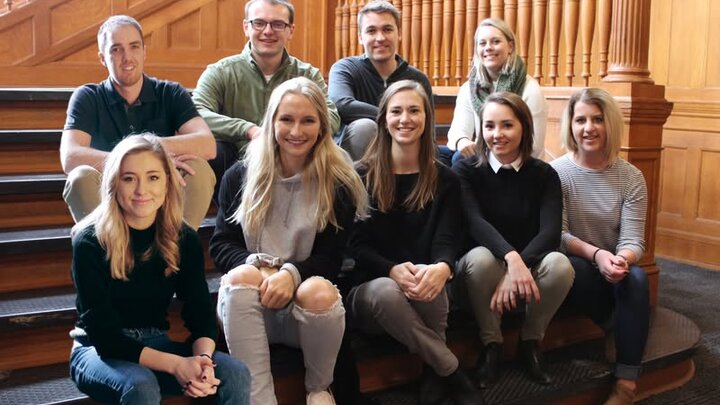 Group sitting on wooden stairs, smiling, in a warm setting with polished railings in the background.