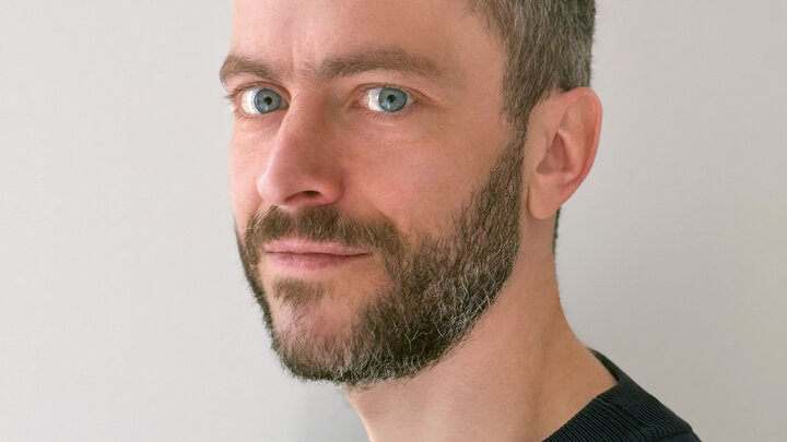 Man with a short beard and gray hair wearing a dark sweater, looking at the camera against a white wall.
