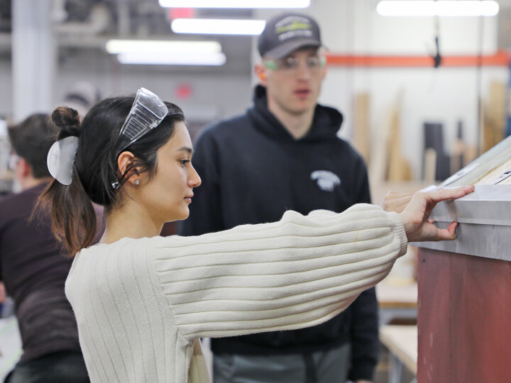 Woman wearing safety goggles operating machinery in a workshop setting; others visible in the background.