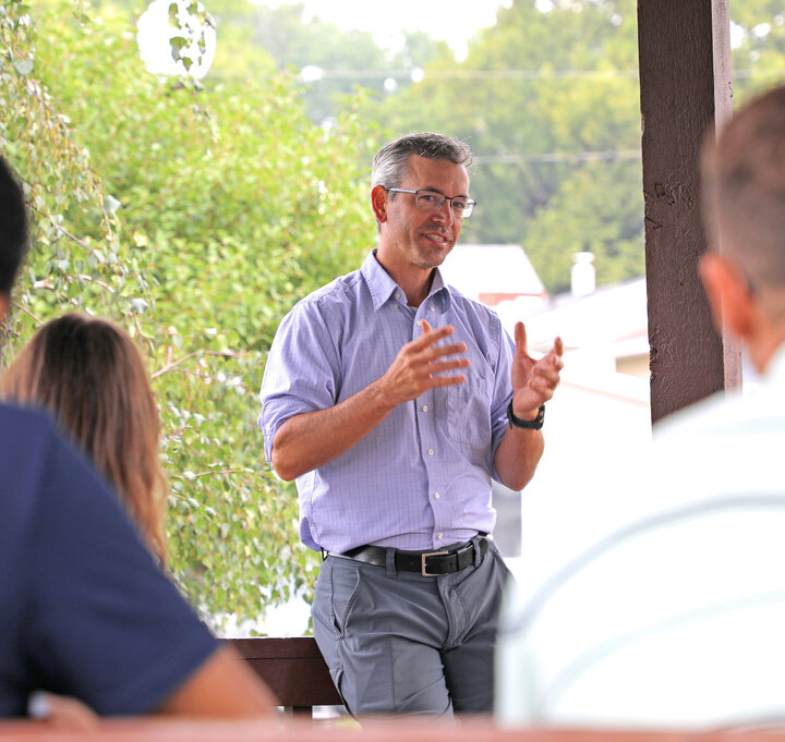 Man gesturing while speaking outdoors to a small group, with greenery in the background.