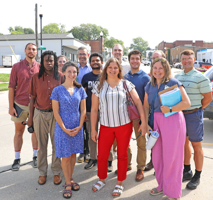 A group of smiling people standing together outdoors on a sunny day in a small town setting.