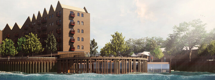 Brick waterfront building with pointed roof, trees, pier, and calm water under a partly cloudy sky.